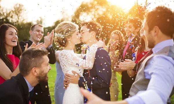 Full length portrait of newlywed couple and their friends at the wedding party showered with confetti in green sunny park