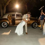 A groom kissing his bride in front of a classic wedding car rental
