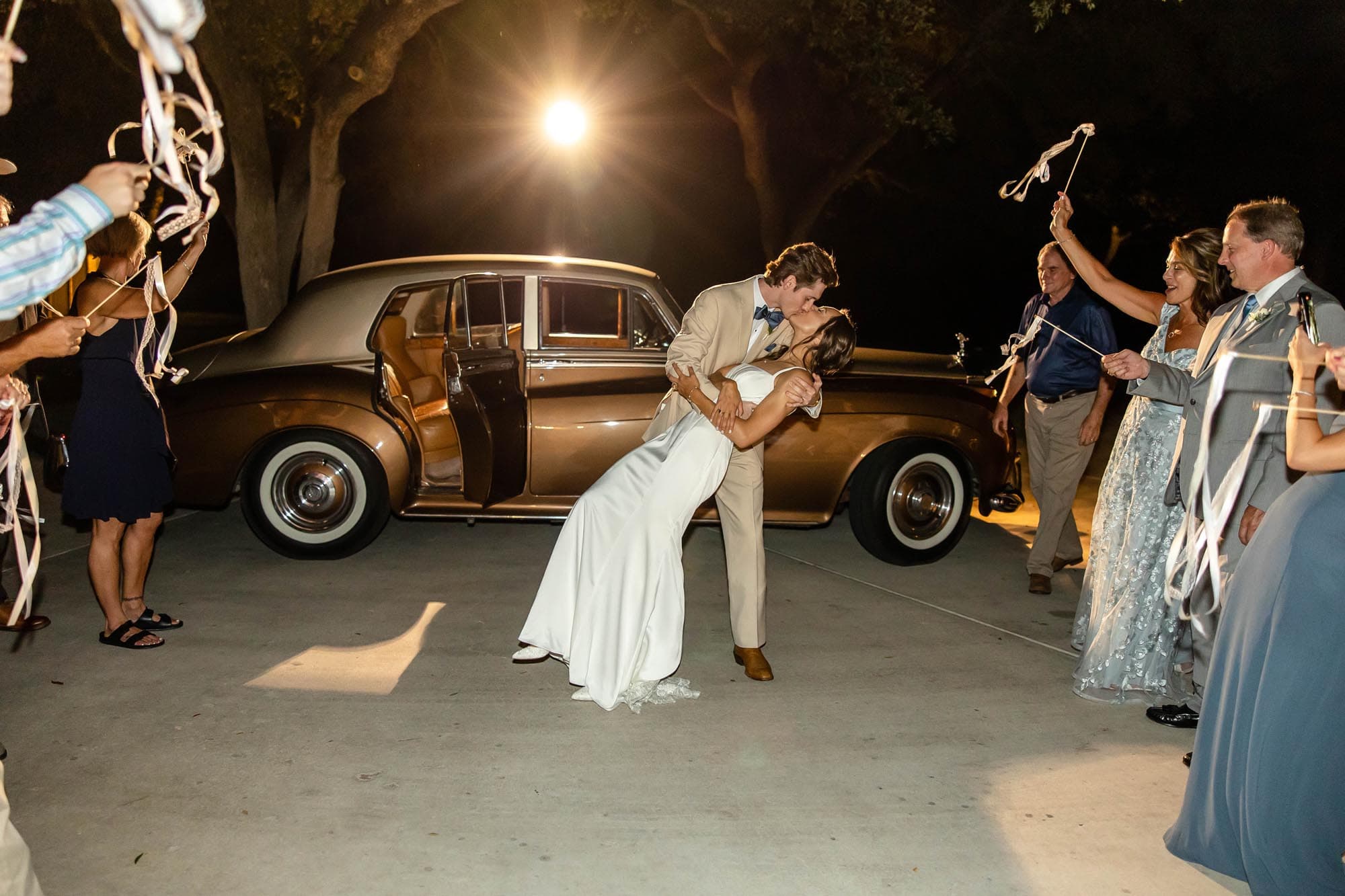 A groom kissing his bride in front of a classic wedding car rental