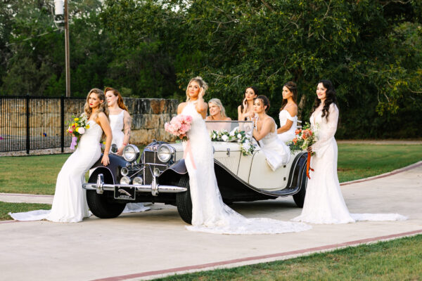 Seven models in white gowns posed around a vintage convertible on a driveway