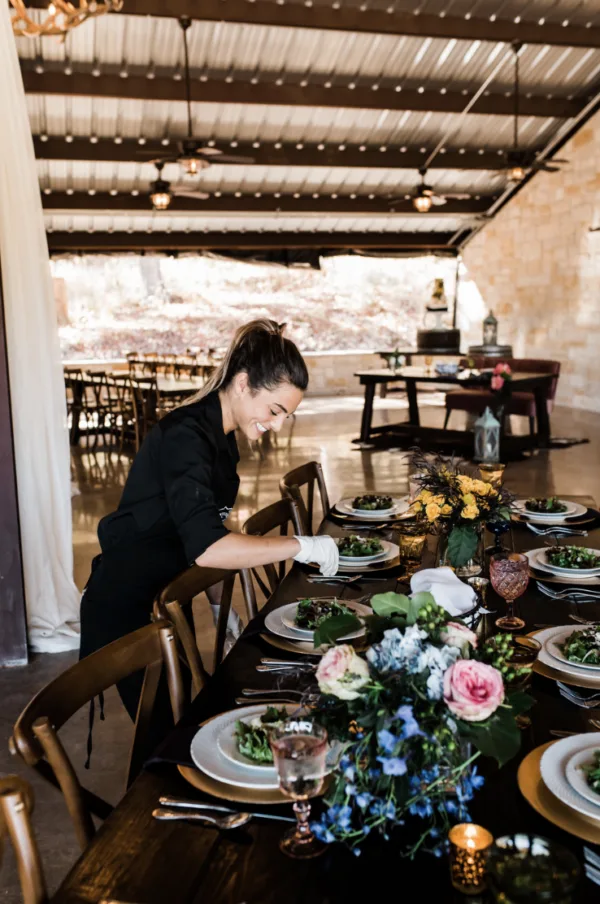 A caterer setting a long wooden table with plated salads, florals and candles