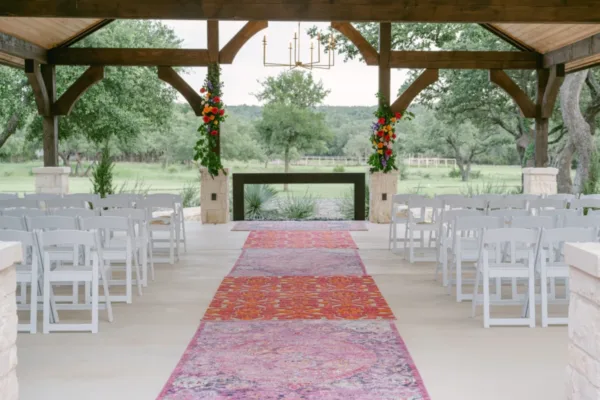A covered outdoor ceremony pavilion with white chairs and a patterned aisle runner
