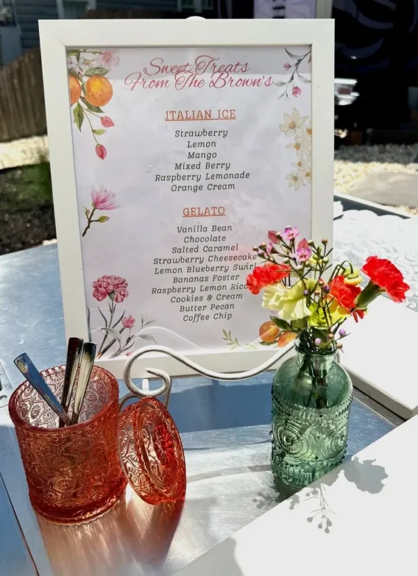 A framed menu listing Italian ice and gelato flavors beside pink glassware and flowers