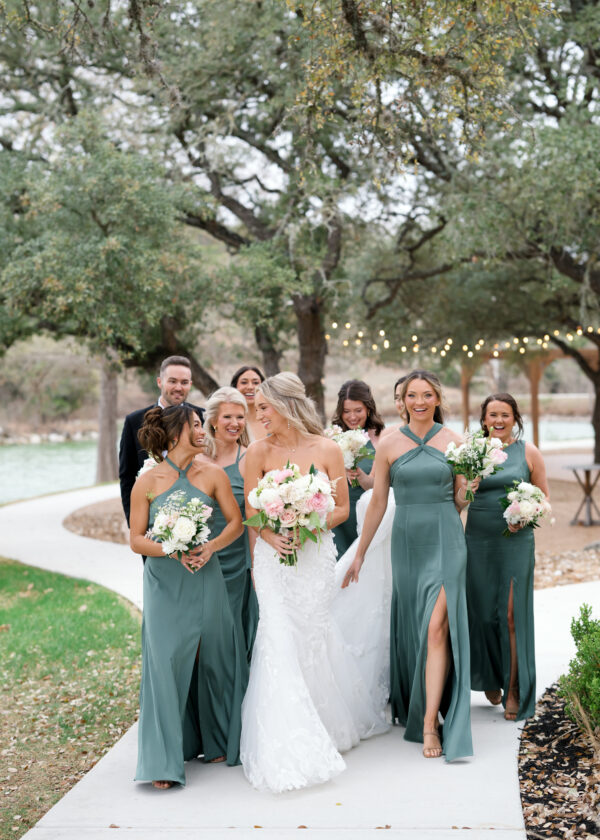 A bride walking with bridesmaids in sage green dresses beneath oaks strung with lights