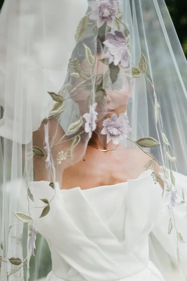 A bride beneath an embroidered floral veil in an off the shoulder gown