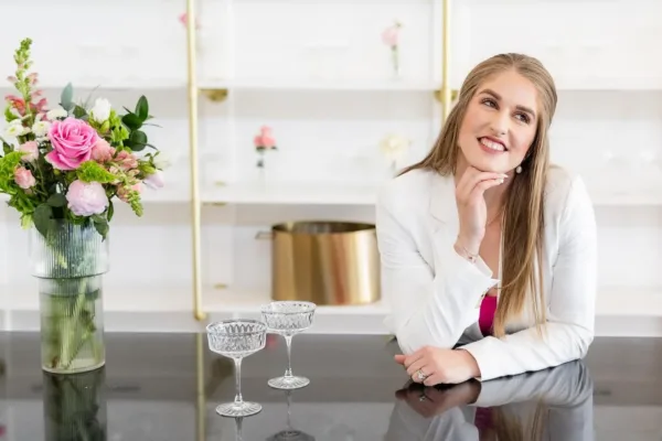 Smiling woman in a white blazer leaning on a bar counter beside coupe glasses and pink roses