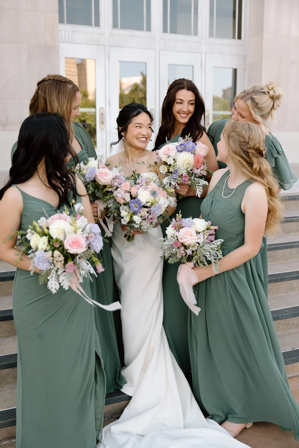 Merribelle laughing with her bridesmaids in sage green dresses holding pastel bouquets