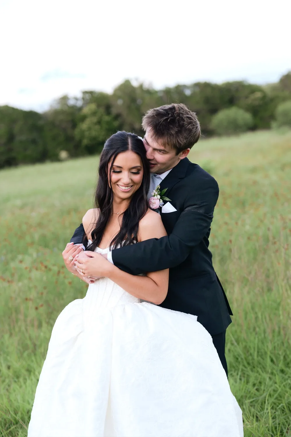 Parker hugs Rebecca from behind in a grassy Texas Hill Country meadow on their wedding day
