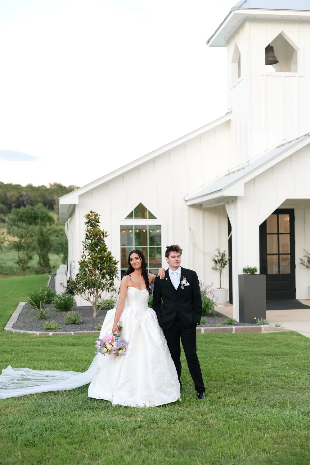 The bride and groom on the lawn before the white chapel at Posey Meadows, her train spread out