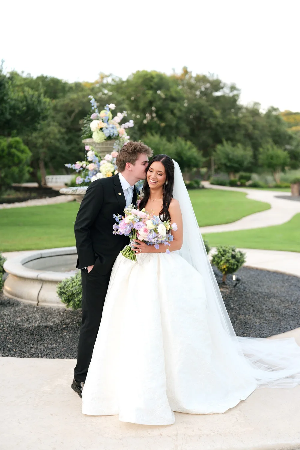 Parker kisses Rebecca on the cheek by the stone fountain, her long veil trailing behind