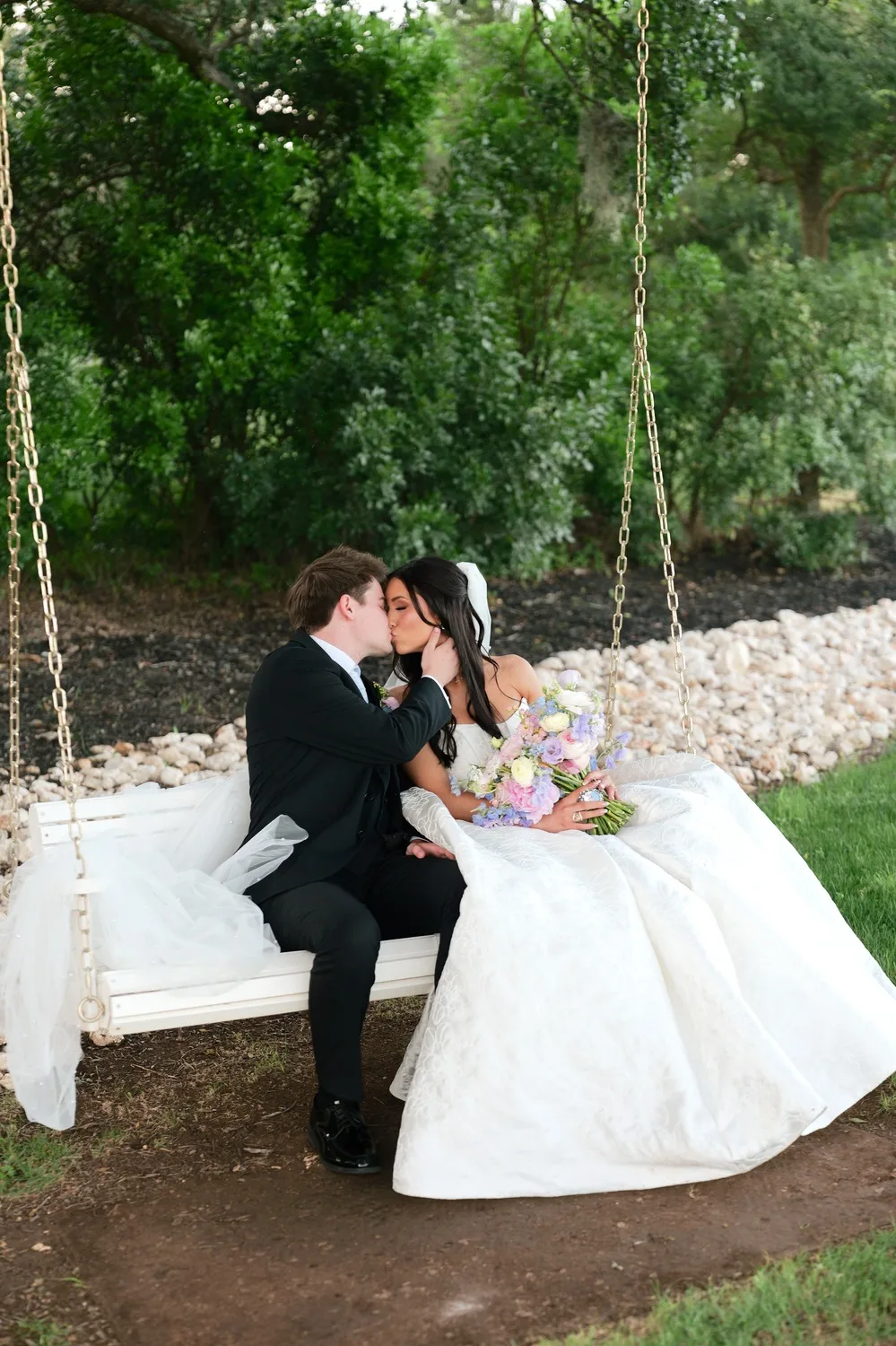 Rebecca and Parker kiss on a white porch swing shaded by trees at Posey Meadows
