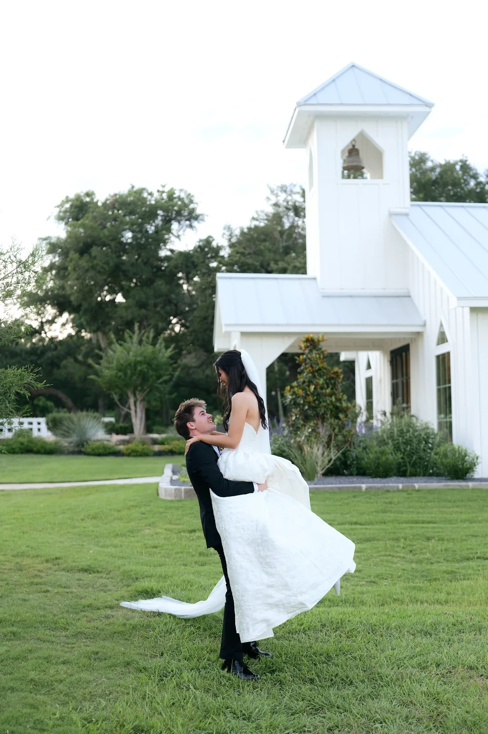 Parker lifts Rebecca off her feet on the lawn in front of the chapel bell tower at Posey Meadows