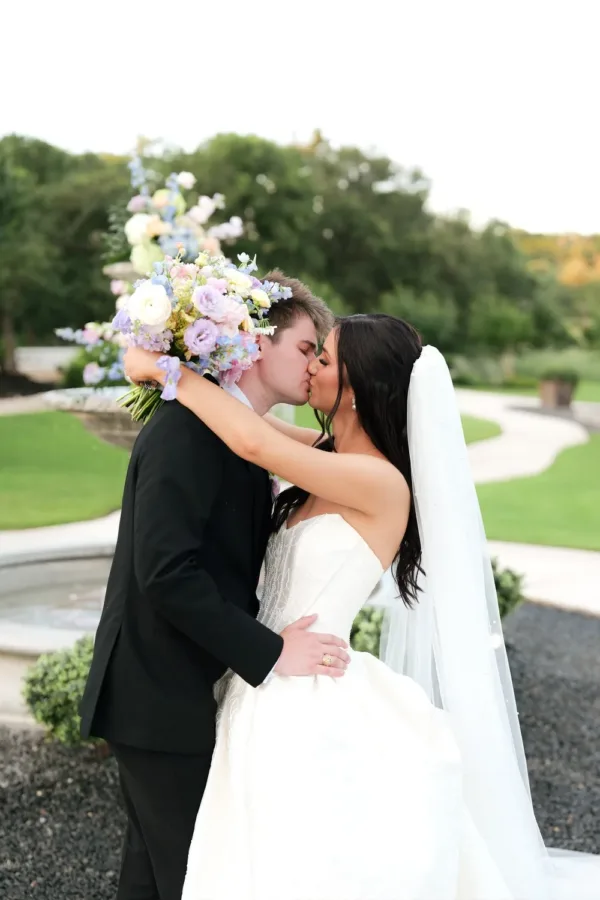 Rebecca and Parker Peterson kiss beside the flower-topped fountain at Posey Meadows in the Texas Hill Country