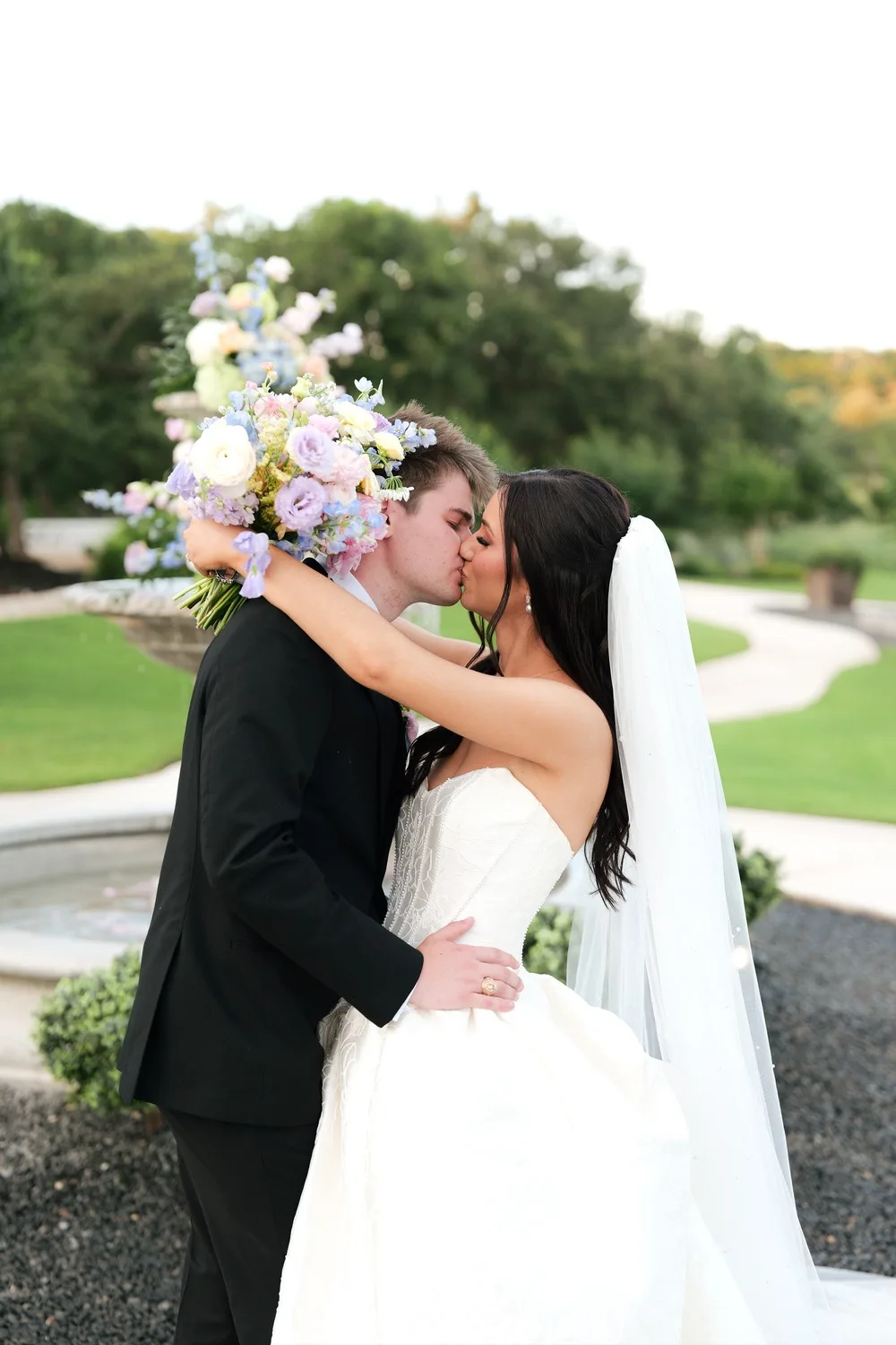 Rebecca and Parker Peterson kiss beside the flower-topped fountain at Posey Meadows in the Texas Hill Country