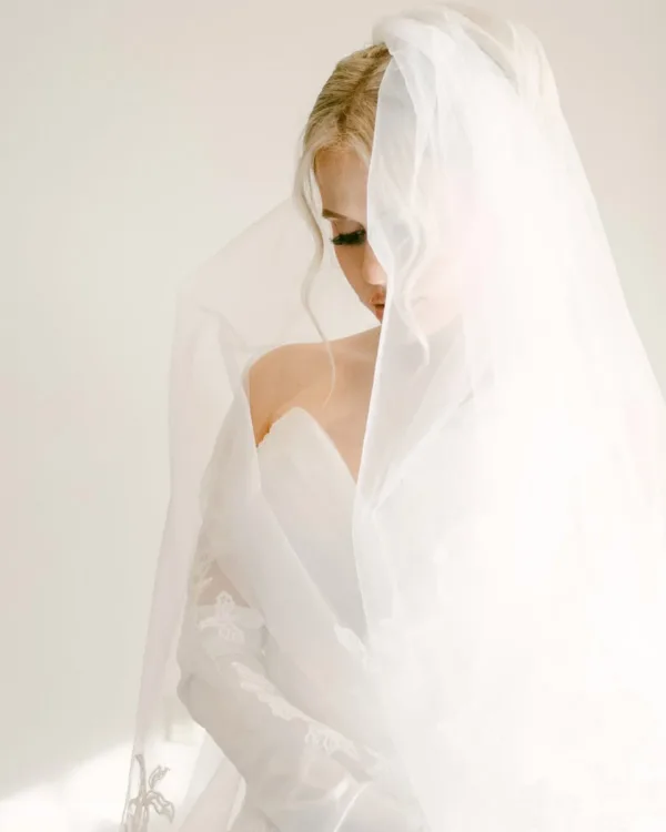 A bride in profile beneath a sheer veil against a pale studio background